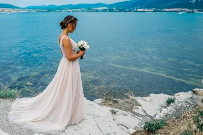 Невеста в платье со шлейфом и букетом в руке на фоне морского пейзажа